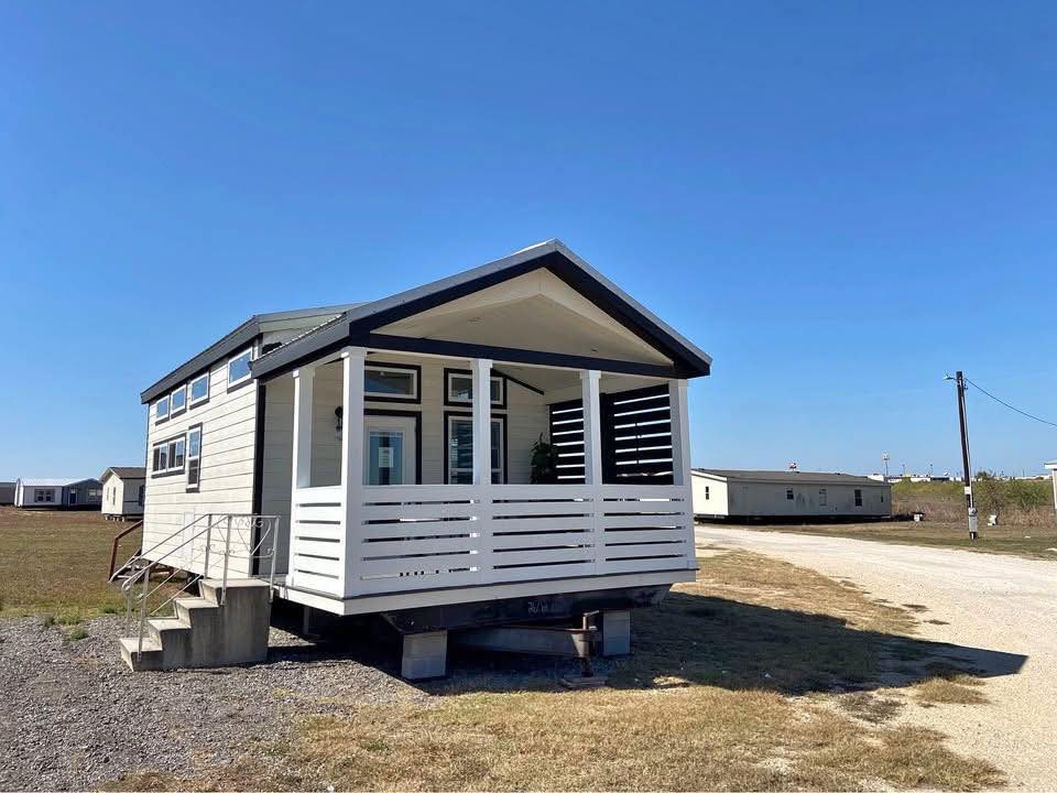 A tiny house with a white exterior and a porch stands on concrete blocks in an open field under a clear blue sky. A dirt road runs alongside it.