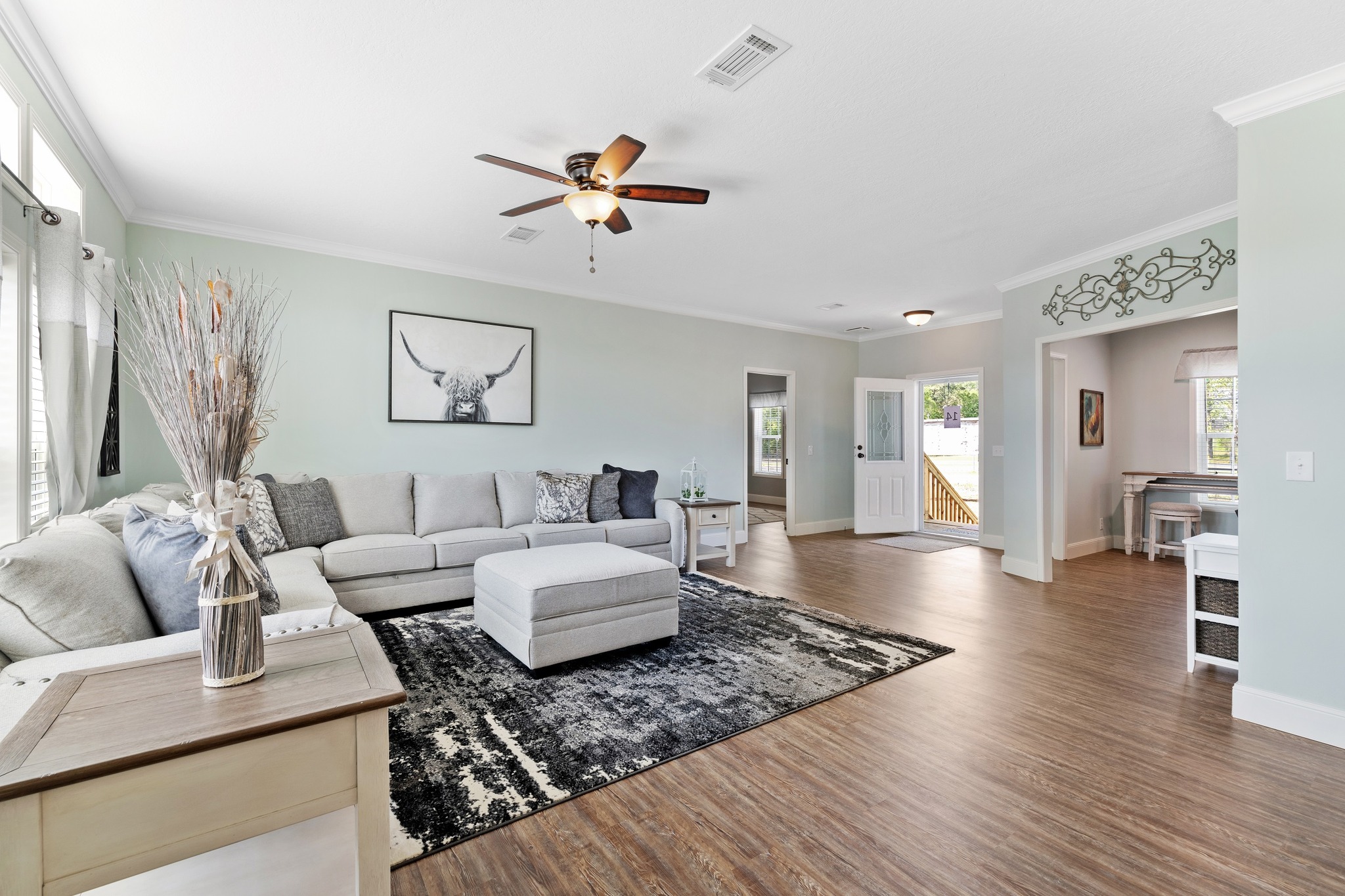 Spacious living room with light green walls and wooden floor, featuring a light gray sectional sofa, abstract art, a ceiling fan, and ample natural light.