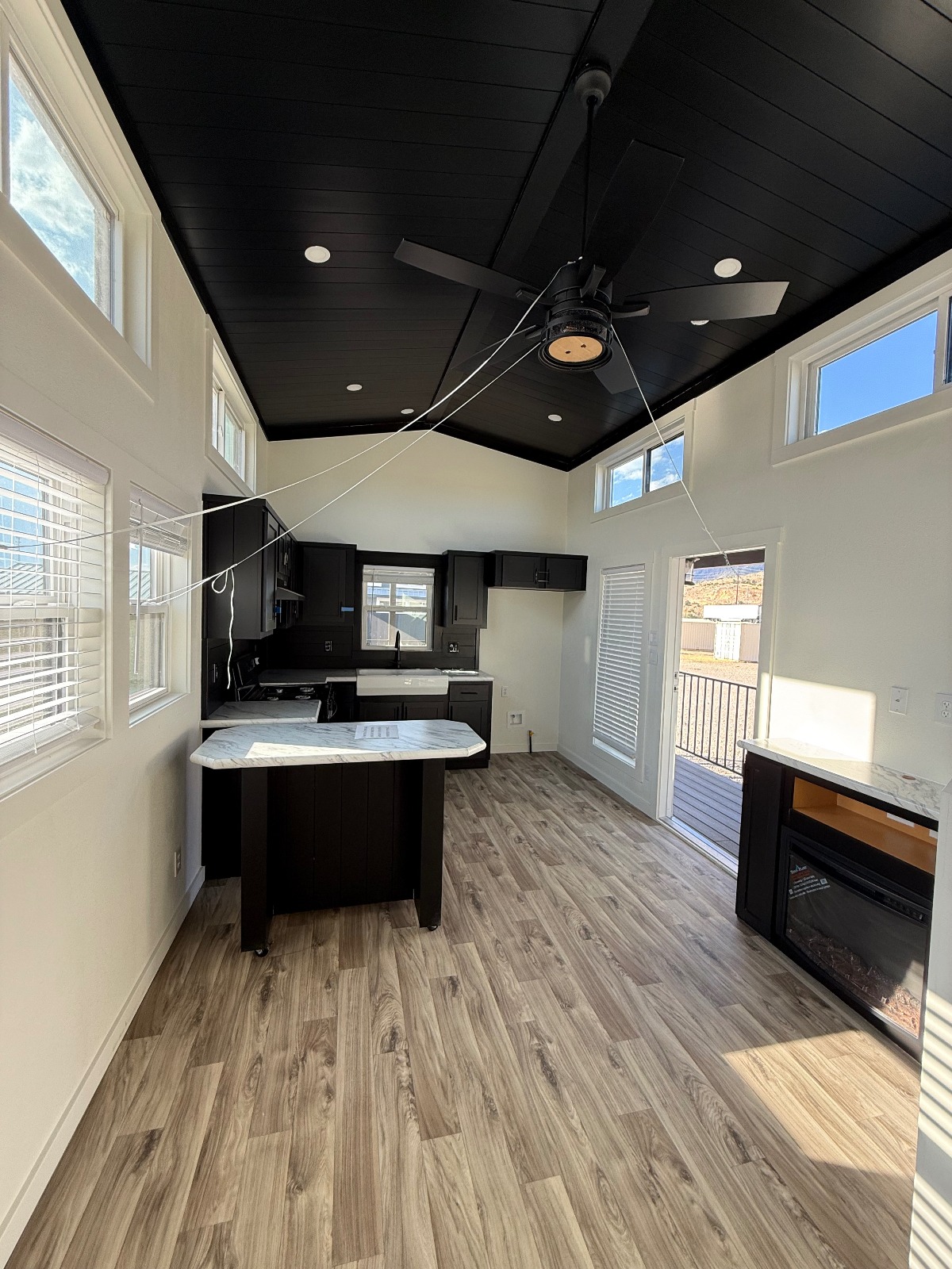 Modern kitchen interior with high ceiling, dark wooden cabinets, center island, and a ceiling fan. Large windows and sliding door provide ample light.