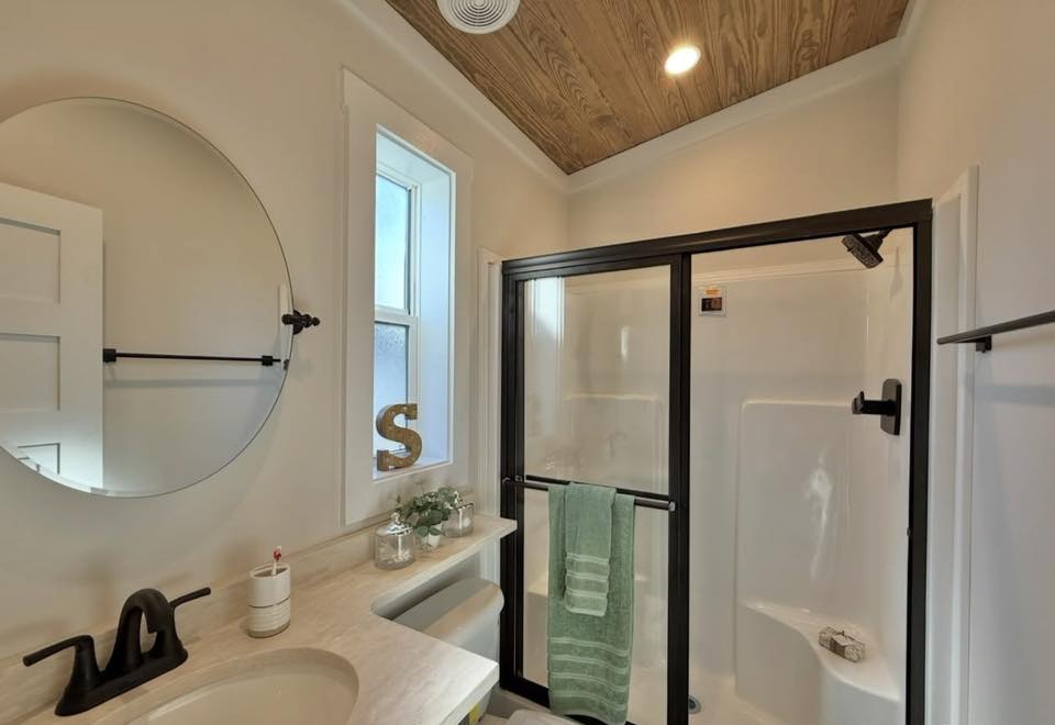 Modern bathroom with a wood-paneled ceiling, black shower frame, round mirror, and minimalist decor. A green towel hangs on the shower door.