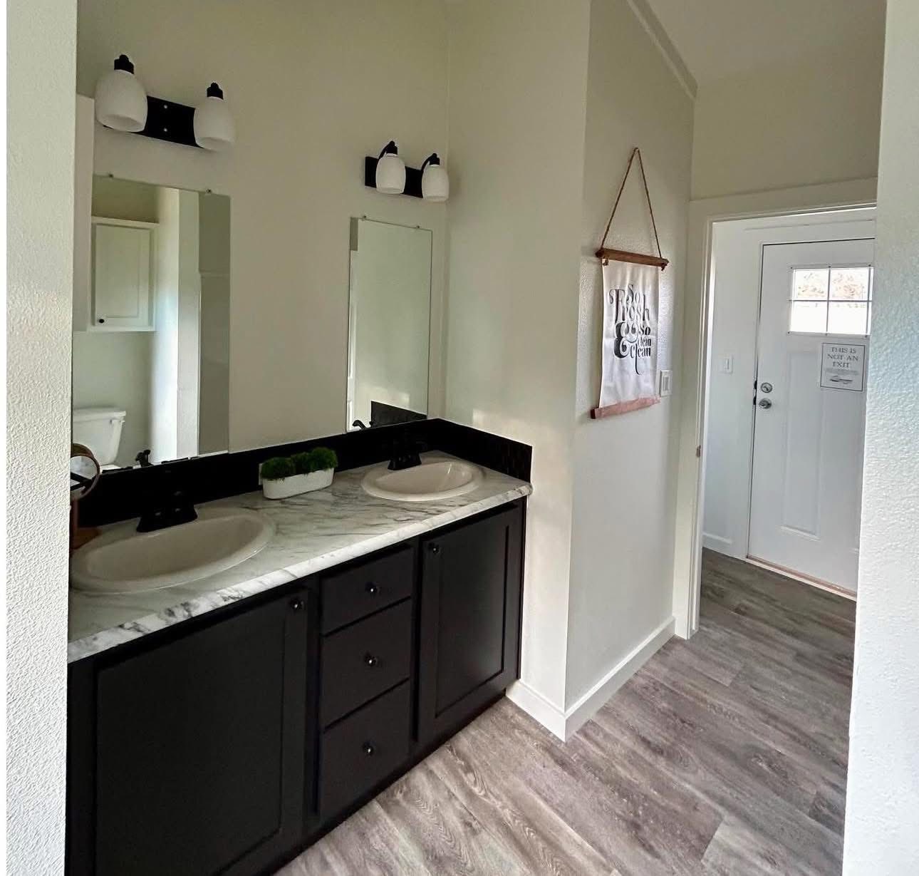 Modern bathroom with dual sinks on a marble countertop and black cabinetry. Bright lighting, minimalist decor, and a door in the background convey a clean, inviting atmosphere.