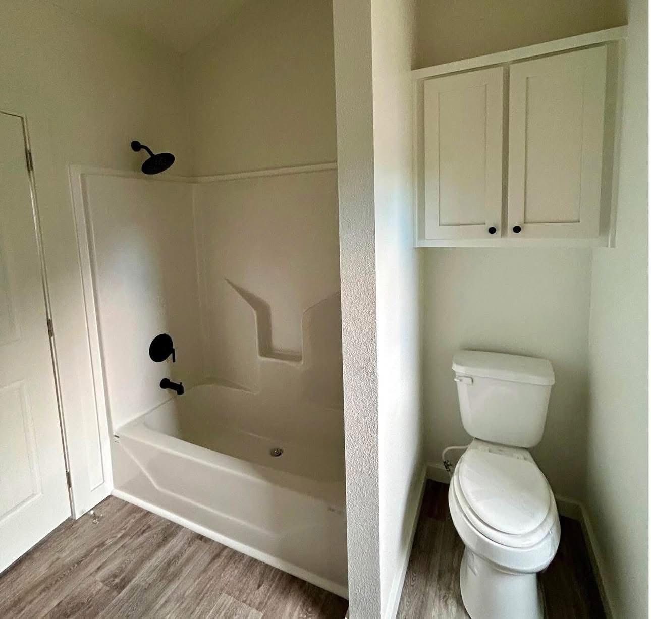 A small bathroom with a white bathtub and shower on the left, a toilet on the right, and a white cabinet above the toilet. The floor is wood-paneled.
