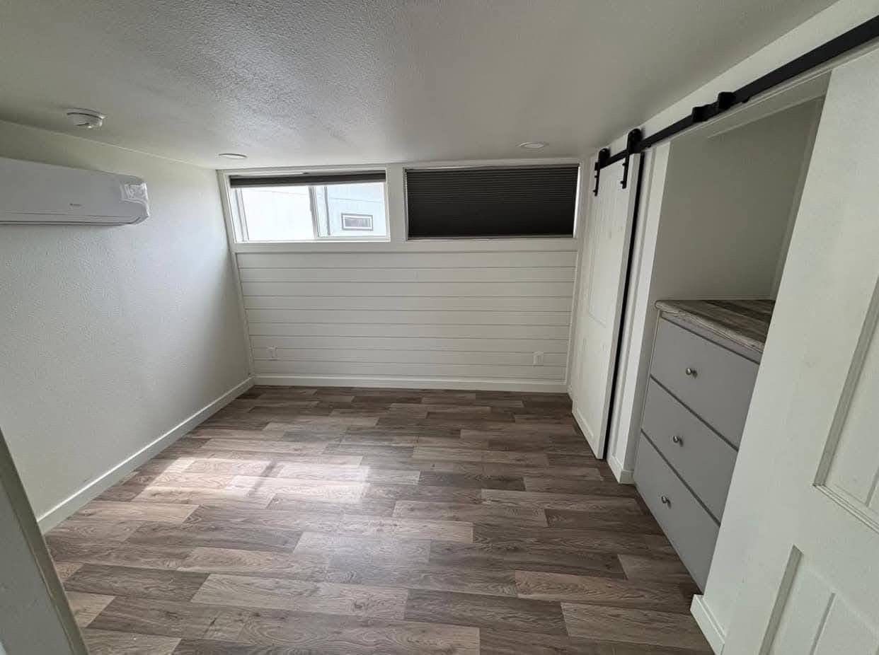 A small, bright room with light wood flooring, a high narrow window, white walls, and a sliding barn door revealing built-in drawers, conveying simplicity.