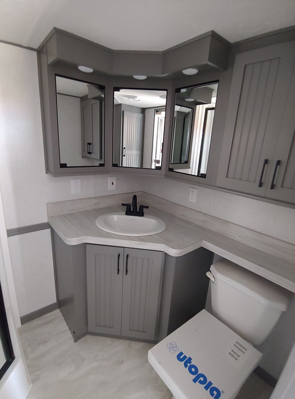 Compact bathroom with gray cabinets and a sleek countertop. The vanity includes a white sink with a black faucet. A toilet with a box labeled "Utopia" is visible. Mirrors reflect the minimalist design, creating a clean and modern feel.