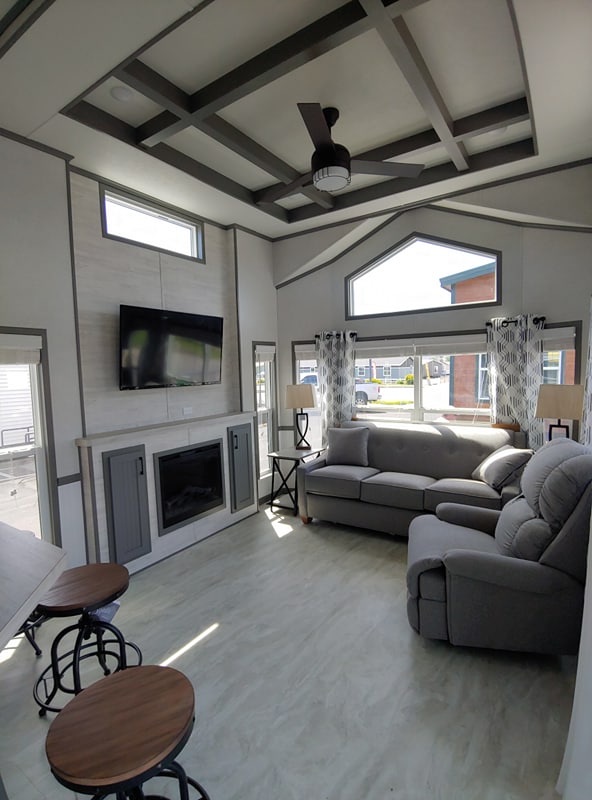 Modern living room with high ceiling and geometric beams, featuring a gray sectional sofa and recliner, wall-mounted TV, fireplace, and large windows. Bright, airy ambiance.