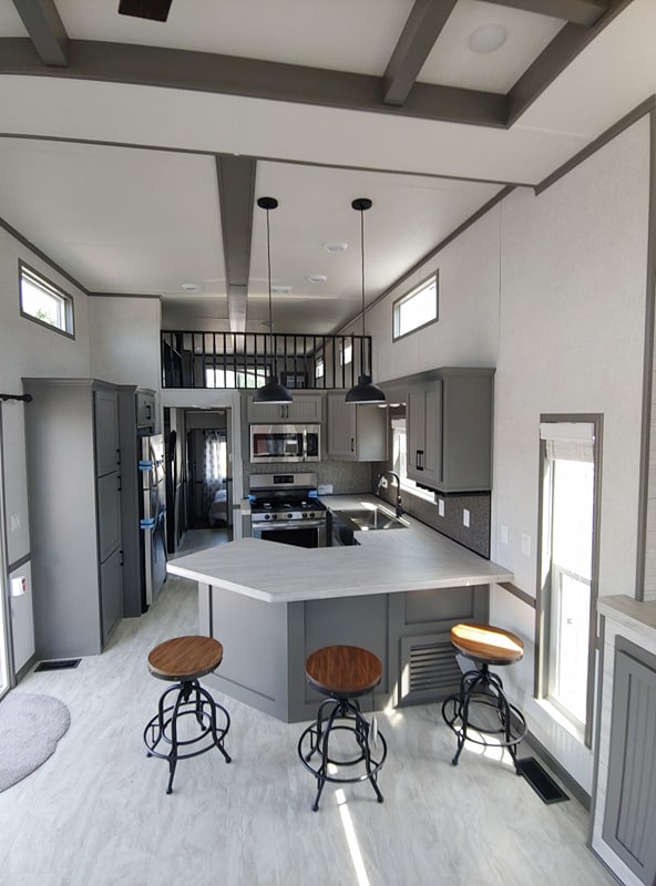A modern kitchen and living area with high ceilings and a loft. Features gray cabinets, stainless steel appliances, and three bar stools by a white island.