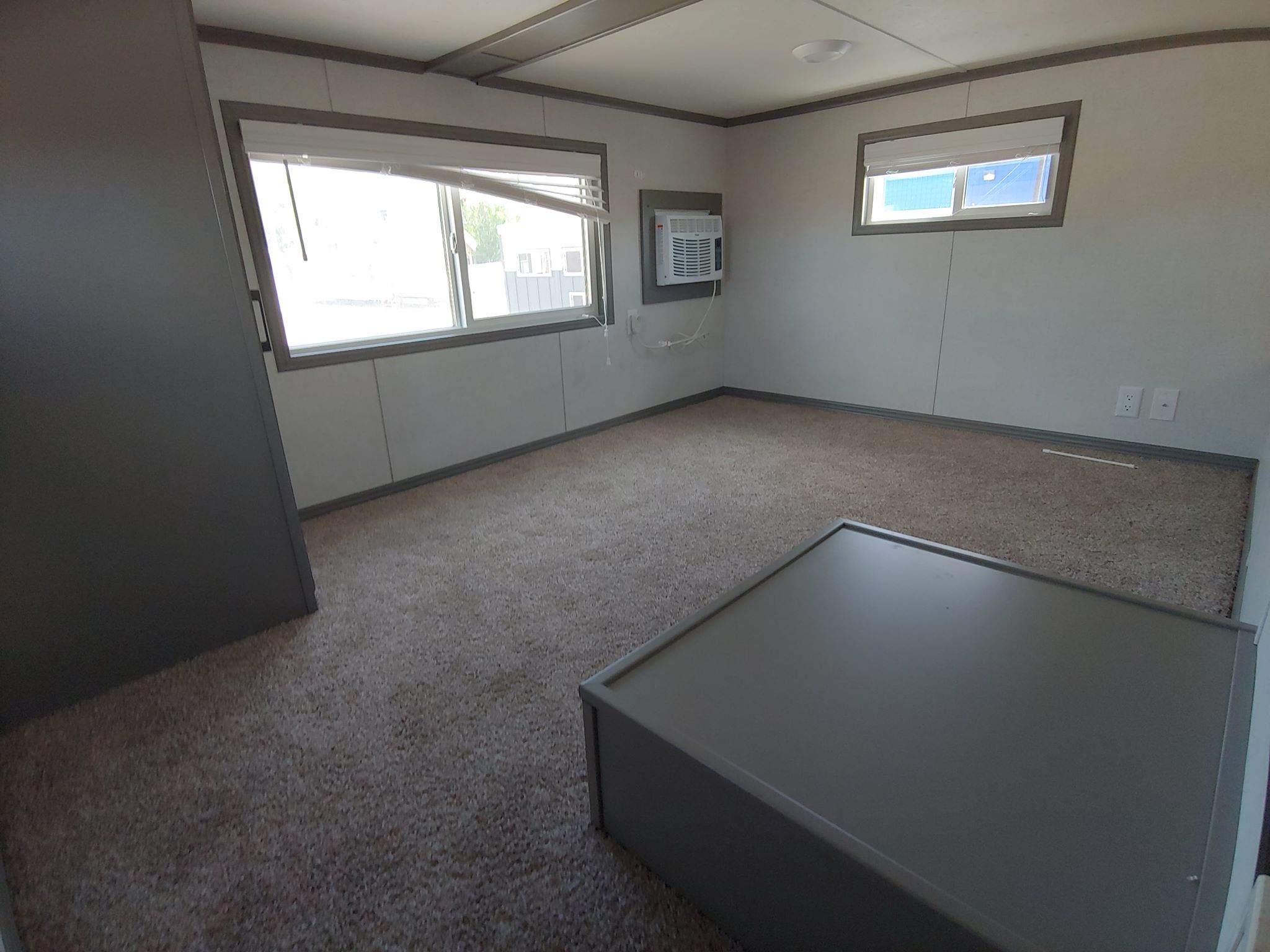 Small, carpeted room with two windows, an air conditioner, and minimalist gray furniture. The space feels clean and simple, with ample natural light.