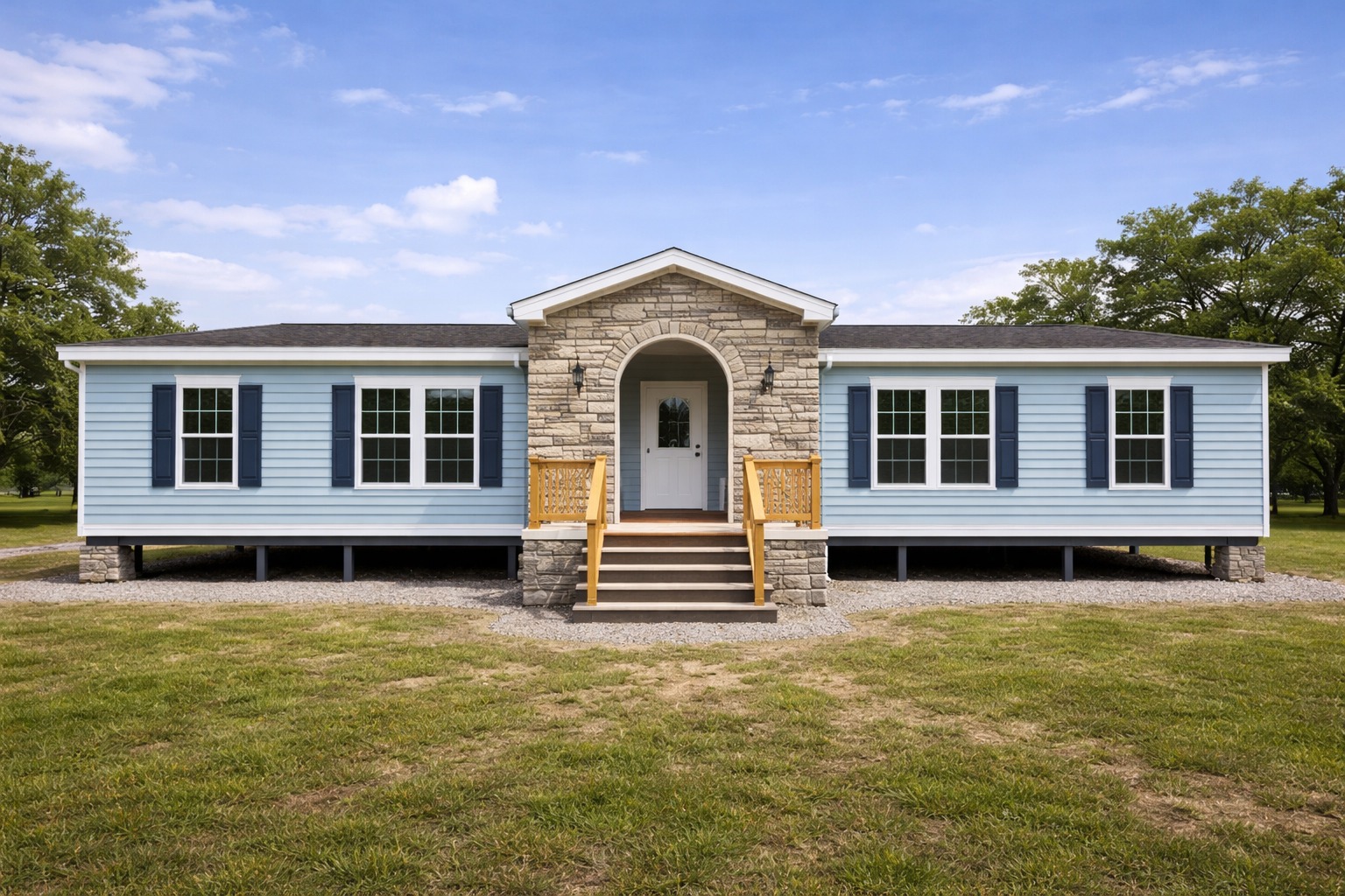A light blue modular home with a central stone entryway and steps. It has white-framed windows and dark shutters, set on a grassy field under a blue sky.