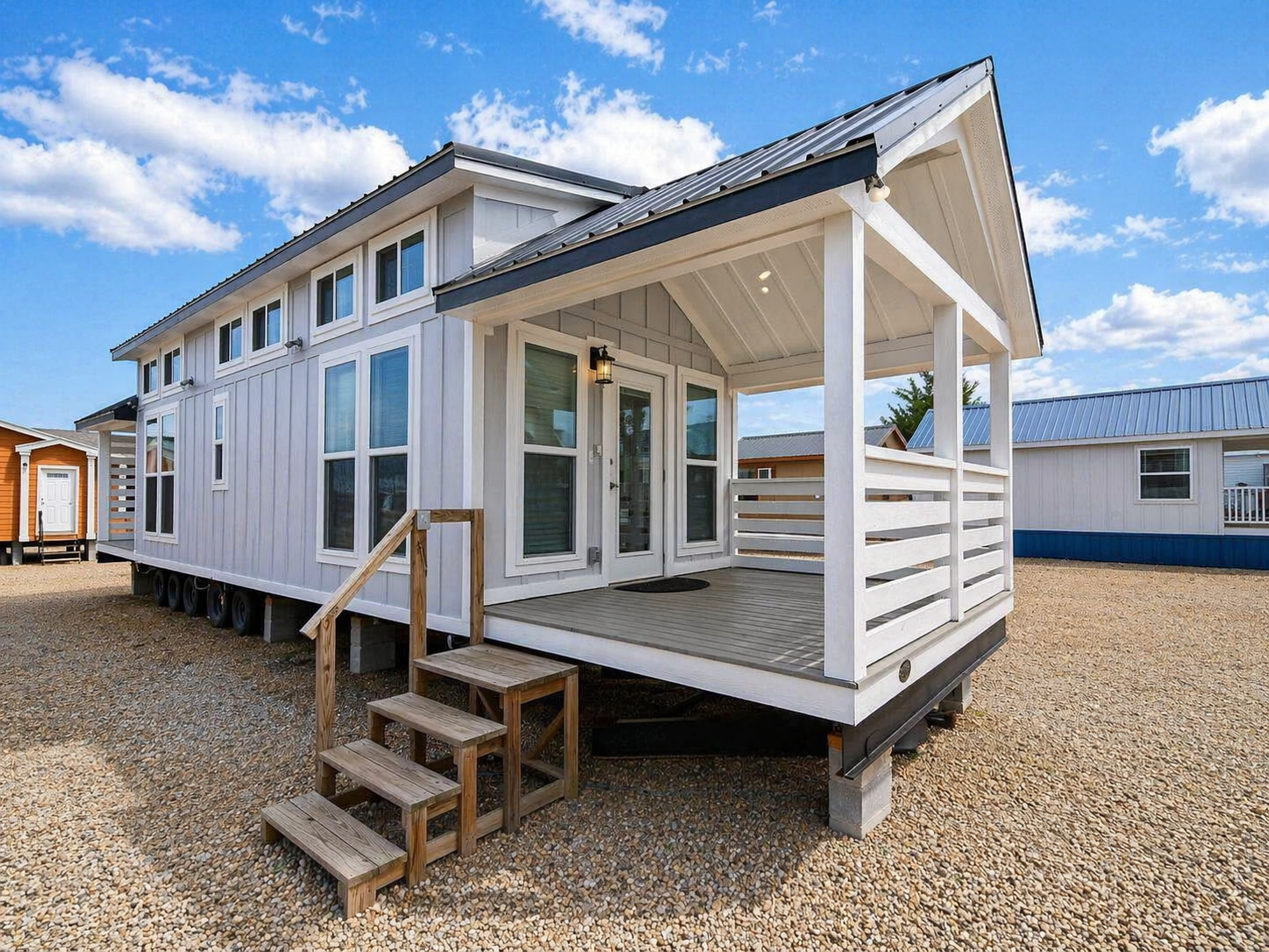 A modern tiny house on wheels with a white exterior, large windows, and a small covered porch. It's set against a clear blue sky and gravel ground.