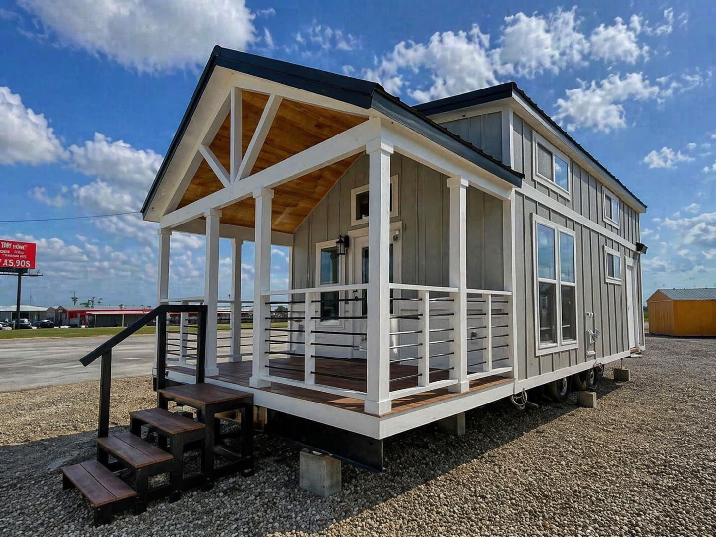 Tiny house on wheels with a porch and white railing, set on a gravel lot under a blue sky. Features large windows and a gabled roof.