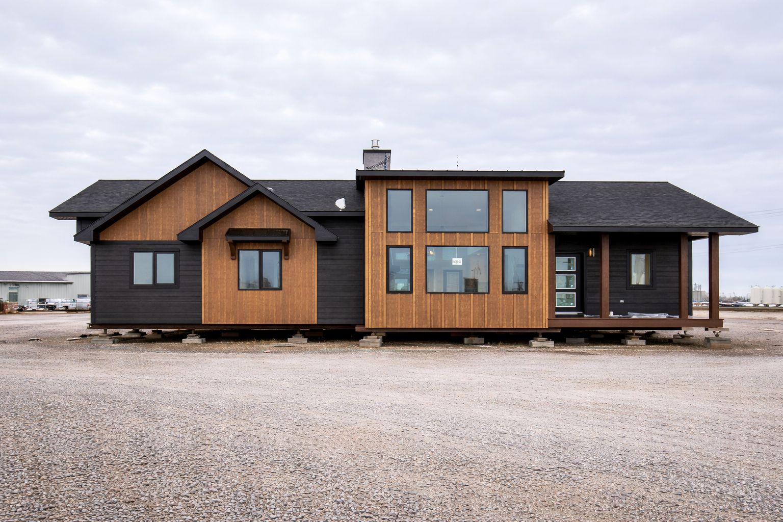 Modern modular home with black and wooden exterior, large windows, flat and gabled roofs, set on a gravel lot under a cloudy sky.