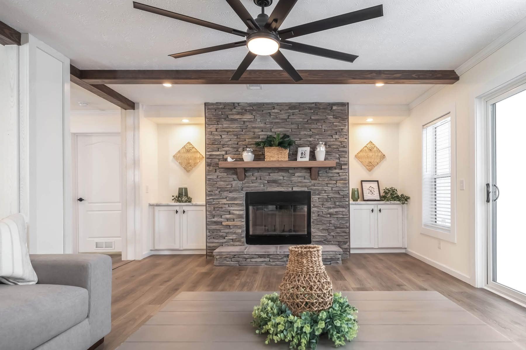 Cozy living room with a stone fireplace, wooden mantel, and ceiling fan. Neutral tones, decorative shelves, and a large window contribute to an inviting atmosphere.