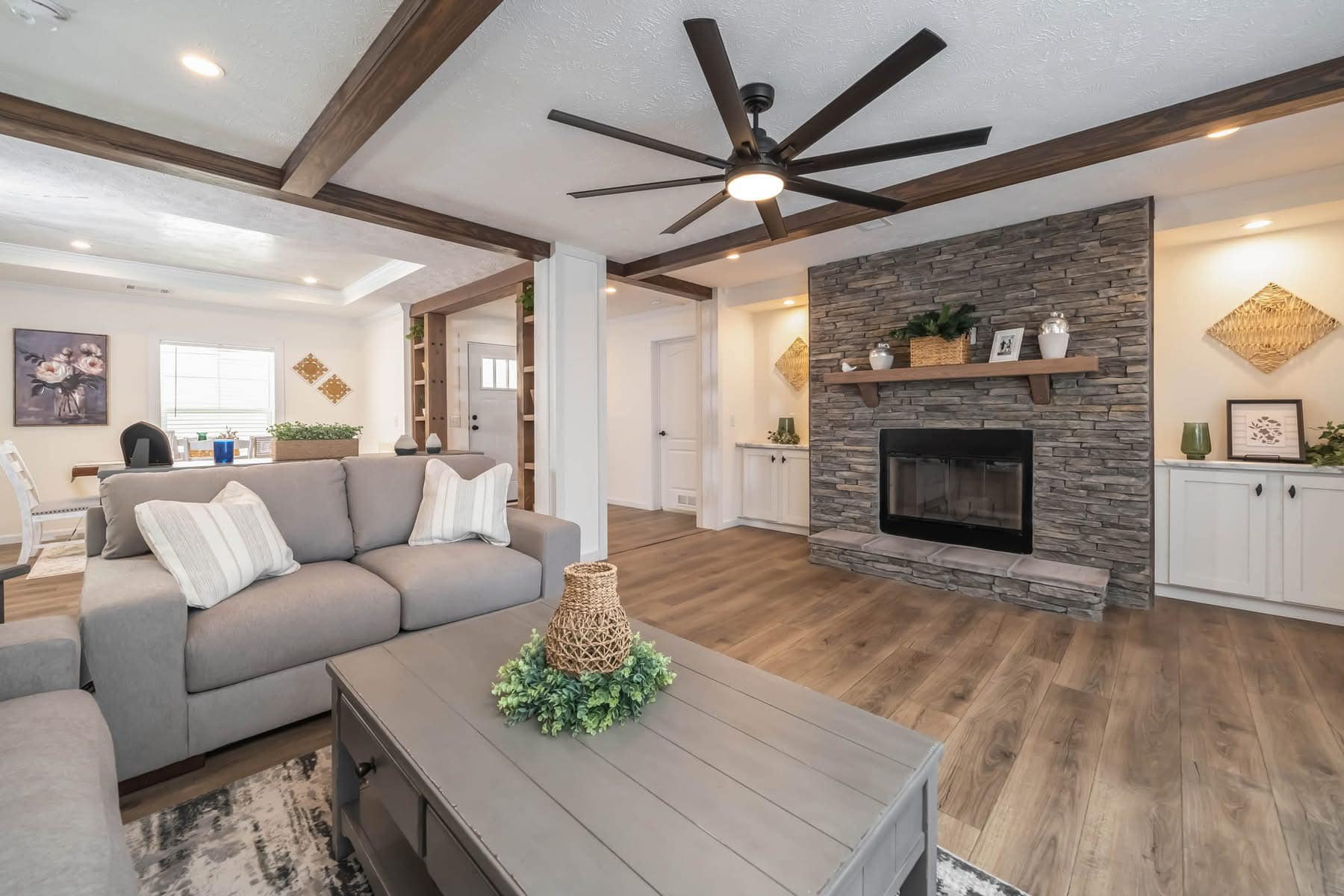 Stylish living room with gray sofas, a large stone fireplace, wooden beams, and a ceiling fan. Cozy, modern decor with green plants on a wooden table.