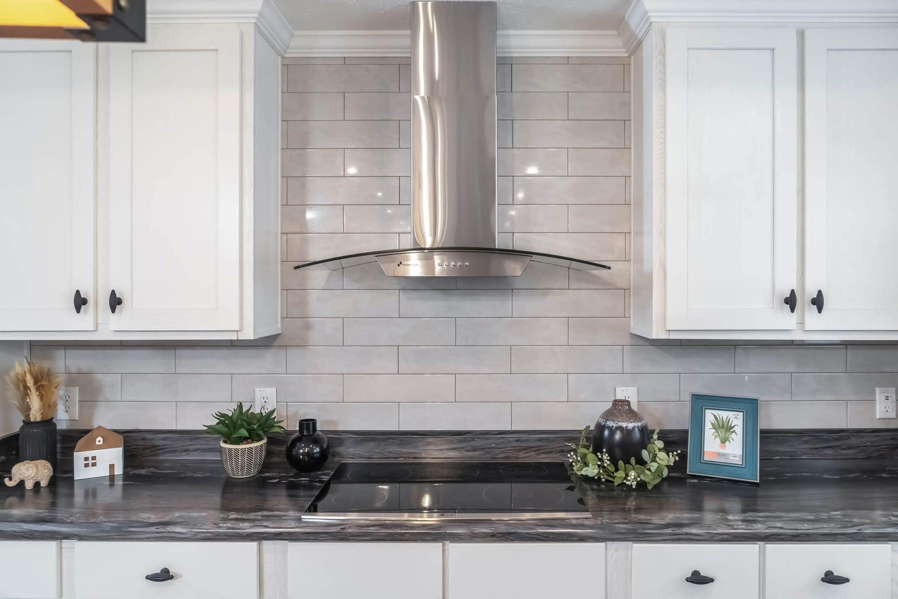 Modern kitchen with white cabinetry, gray subway tile backsplash, and a stainless steel range hood. Decor includes plants and decorative objects.