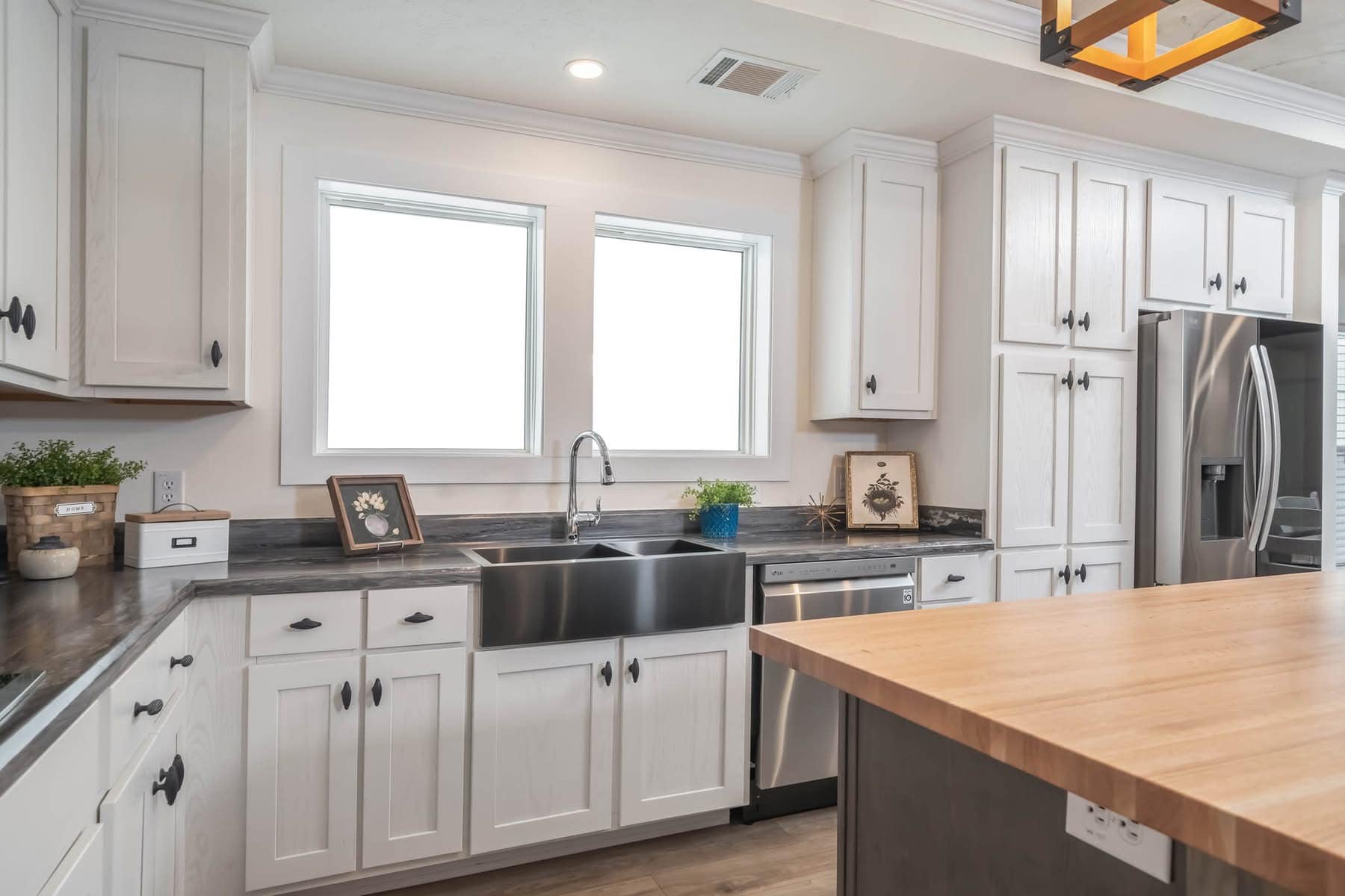 Spacious kitchen with white cabinets, stainless steel sink, gray countertops, and a wooden island. Bright ambiance with framed decor and plants.