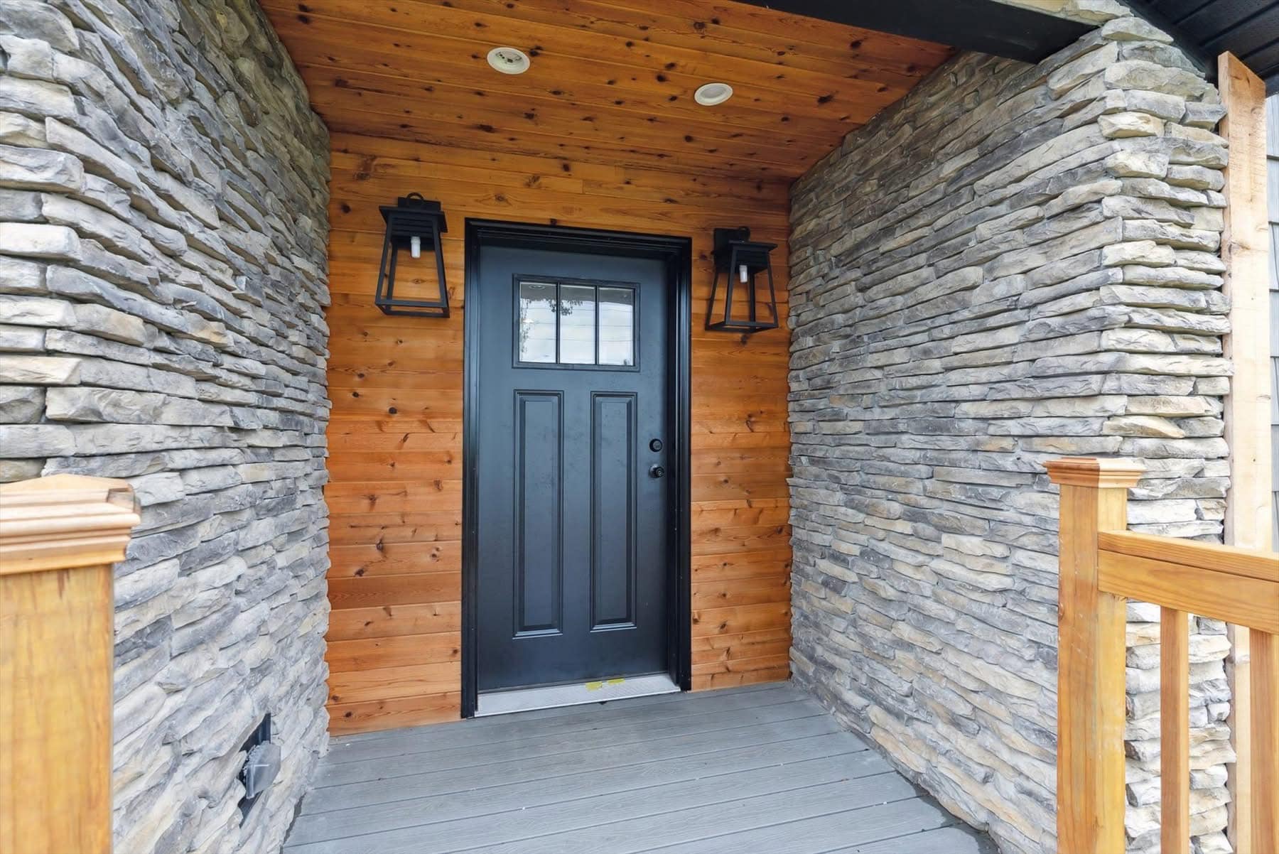 Front porch with a black door framed by wooden siding and stone walls, flanked by modern lanterns. The space feels rustic and inviting.