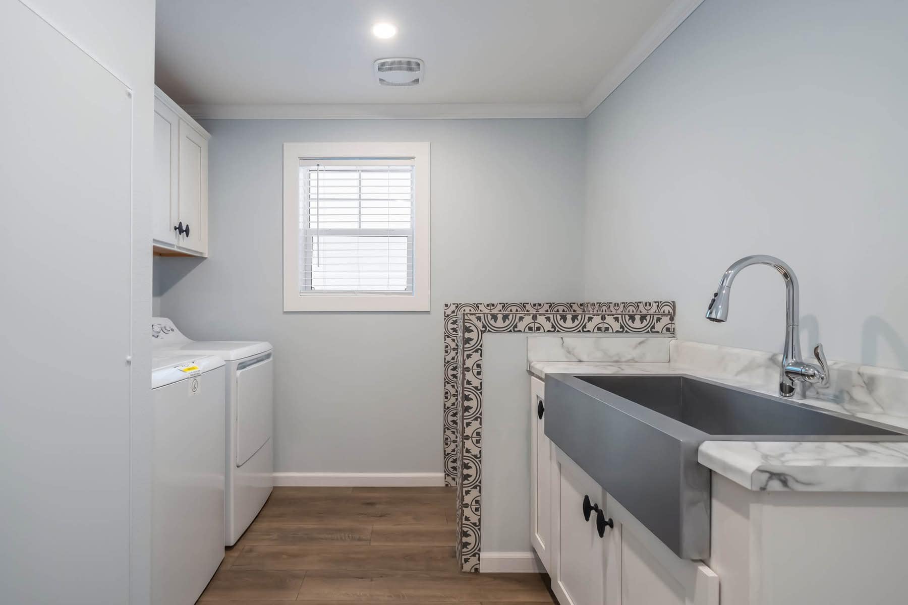 Modern laundry room with light blue walls, a window, washer and dryer on the left. On the right, a deep sink with marble countertop and patterned backsplash.