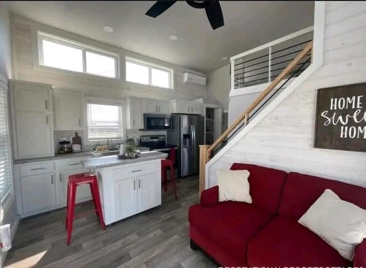 A cozy, modern tiny home interior featuring a red sofa with white pillows, a small kitchen island with red stools, and a "Home Sweet Home" sign by the stairs.