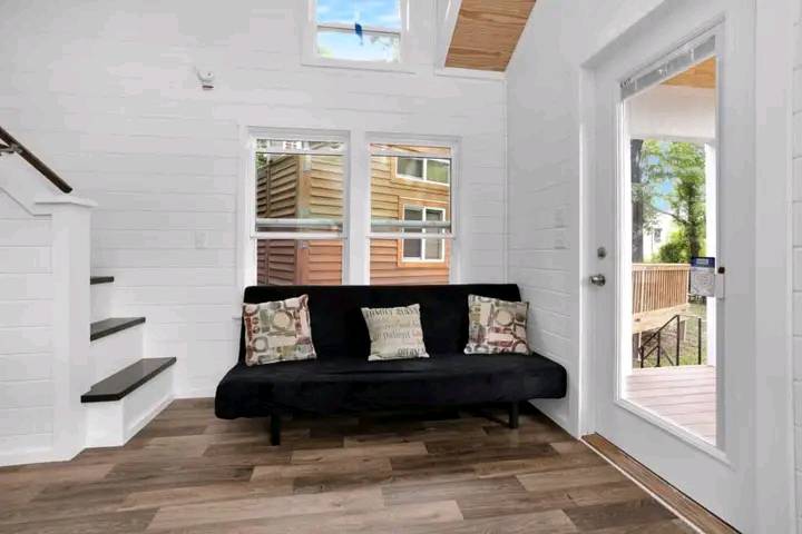 Bright, cozy room with white walls and wood flooring. A black sofa with patterned cushions sits beneath two windows. A glass door leads outside.