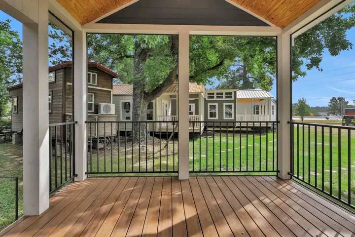 View from a wooden porch with black railing, overlooking a grassy yard and multiple small houses under a tree, creating a peaceful, inviting scene.