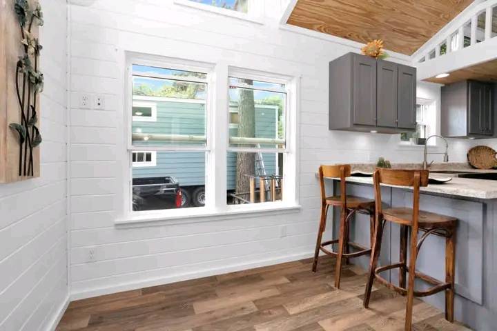 Bright kitchen with wooden bar stools at a counter, white shiplap walls, gray cabinets, and windows overlooking a parked trailer and trees. Cozy and inviting.