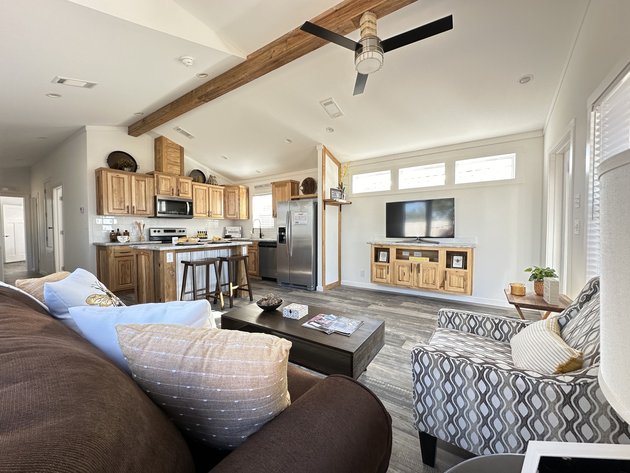 Modern living room with rustic accents. Features a brown sofa, patterned armchair, wood-beam ceiling, and kitchen with wooden cabinets. Cozy and inviting.