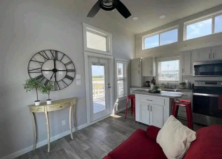 Modern kitchen and living area with natural light. Features a red sofa, wall clock, and bright white cabinets. Red stools add a pop of color. Calm ambiance.