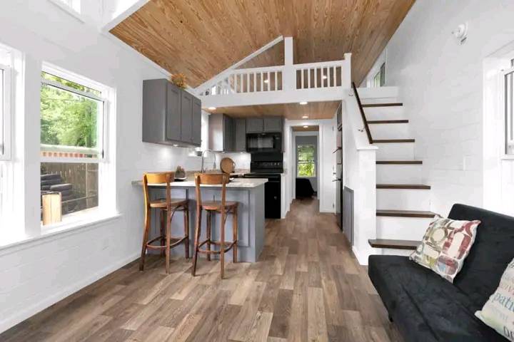 Cozy tiny home interior with a wooden ceiling, loft, and stairs. Features include a kitchen with bar stools, couch with pillows, and large windows.