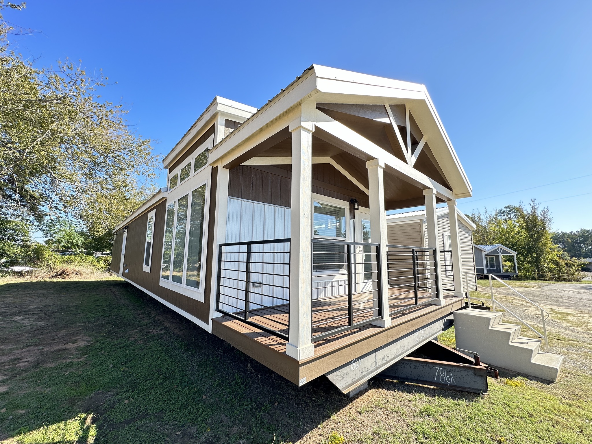 A modern tiny house with a gable roof and wooden porch stands in a grassy area. The bright sky enhances its welcoming, minimalist design.