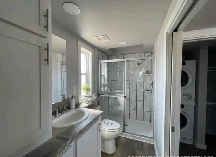 Modern bathroom featuring a sleek gray vanity, white sink, toilet, glass-enclosed shower, and tile accents. Soft lighting; a stacked washer and dryer be seen through an open door.