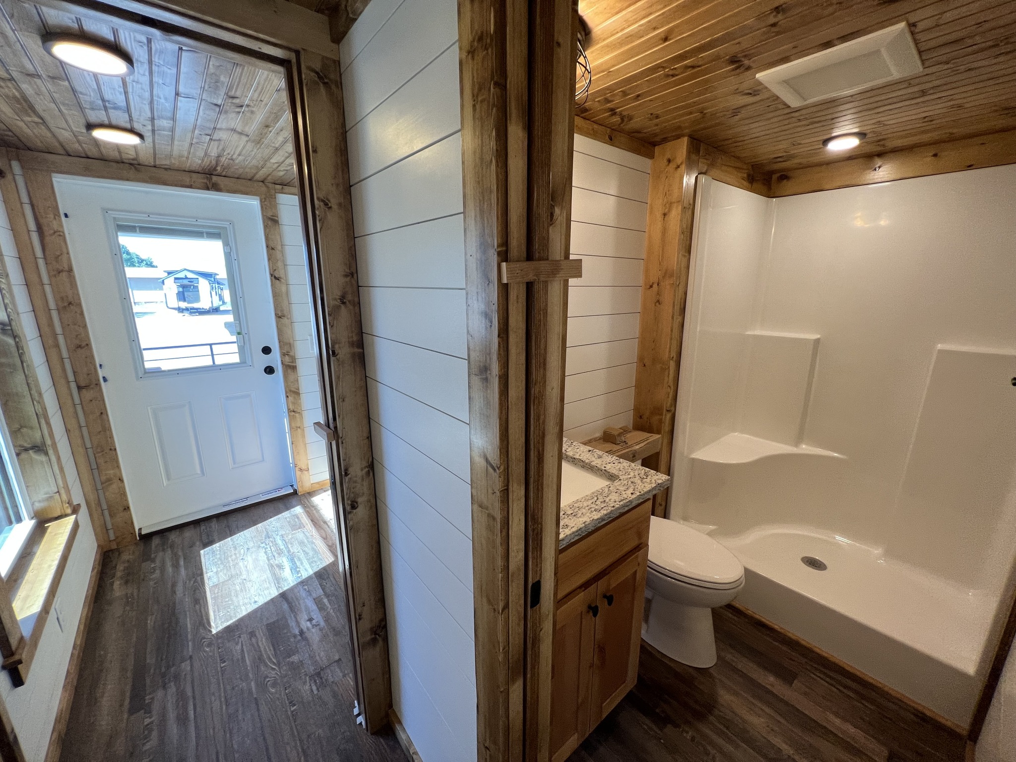 Cozy bathroom with wood accents, white paneled walls, granite countertop, and a shower. Adjacent hallway leads to a door, enhancing natural light.