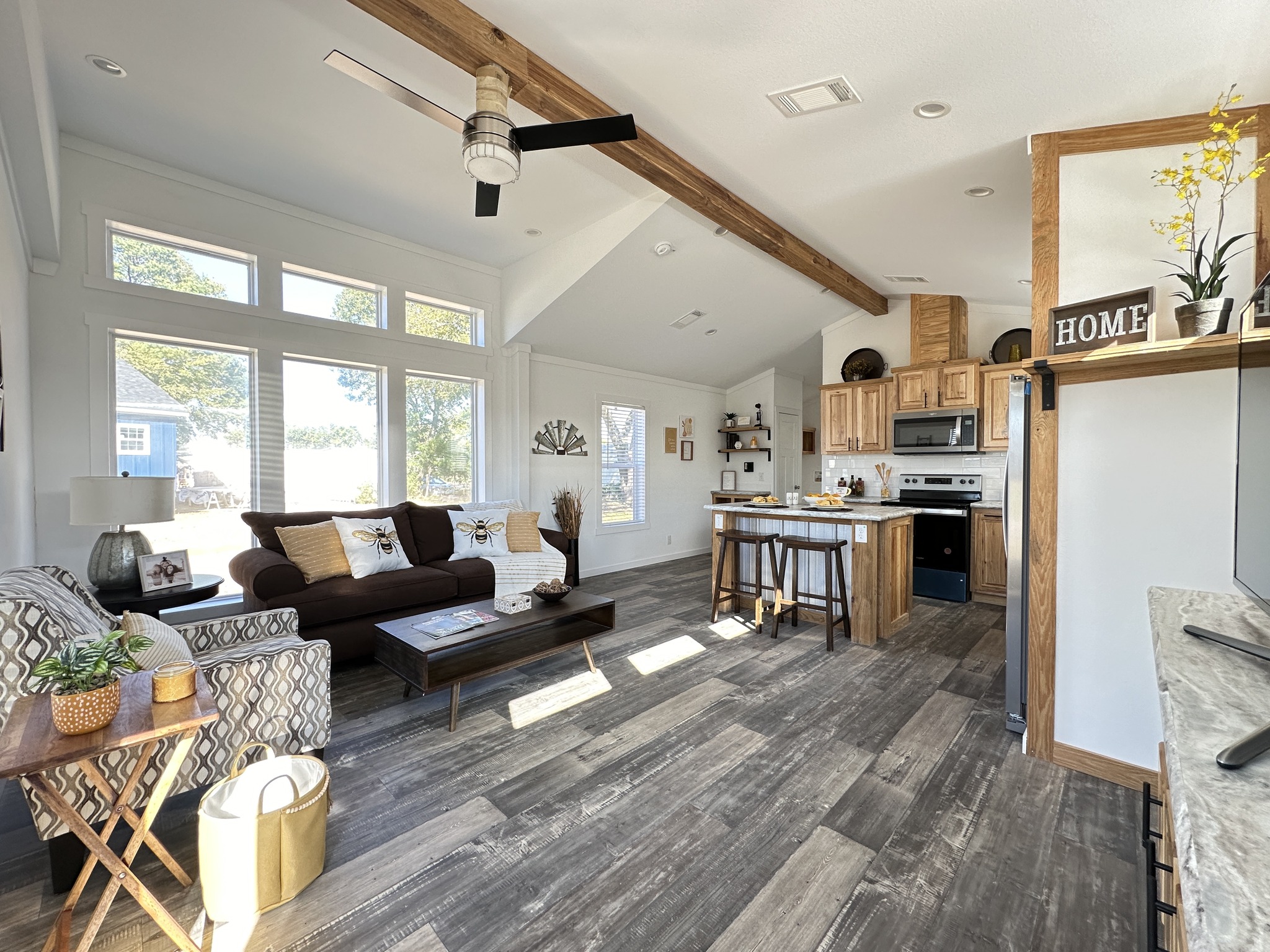 Spacious living room with natural light from large windows. Features a brown sofa, patterned armchair, modern kitchen, and wooden ceiling beam. Cozy and inviting atmosphere.