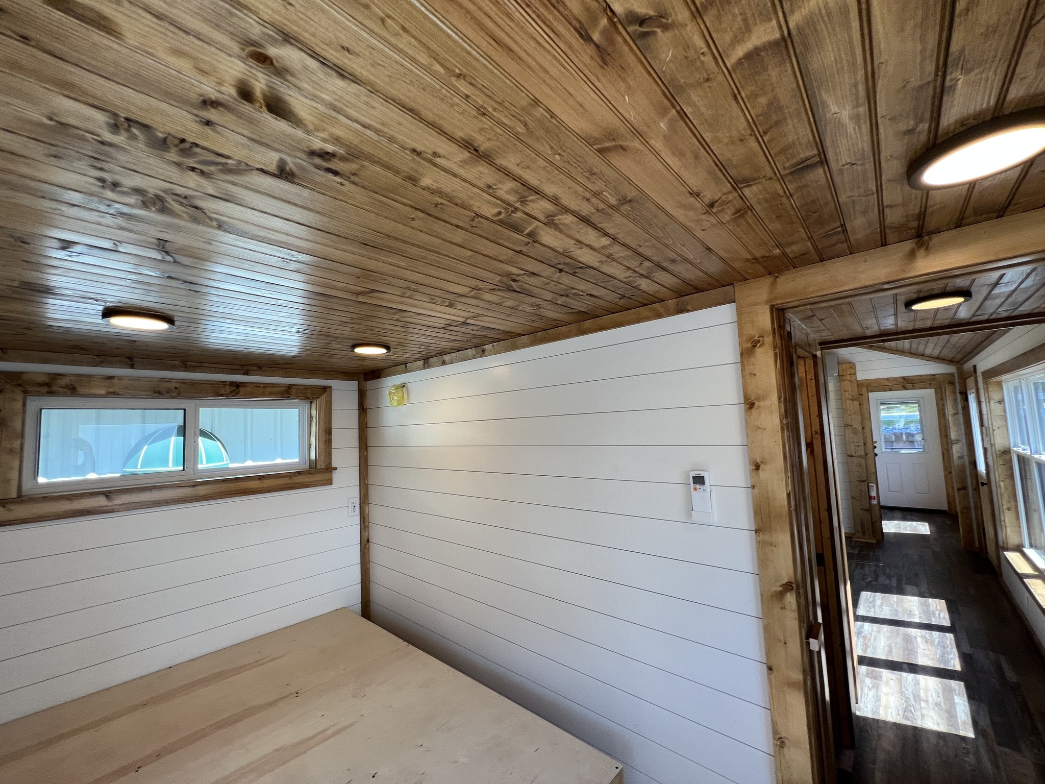 Compact interior of a tiny home with wooden ceiling and white shiplap walls. Soft lighting and narrow windows enhance the cozy, rustic ambiance.