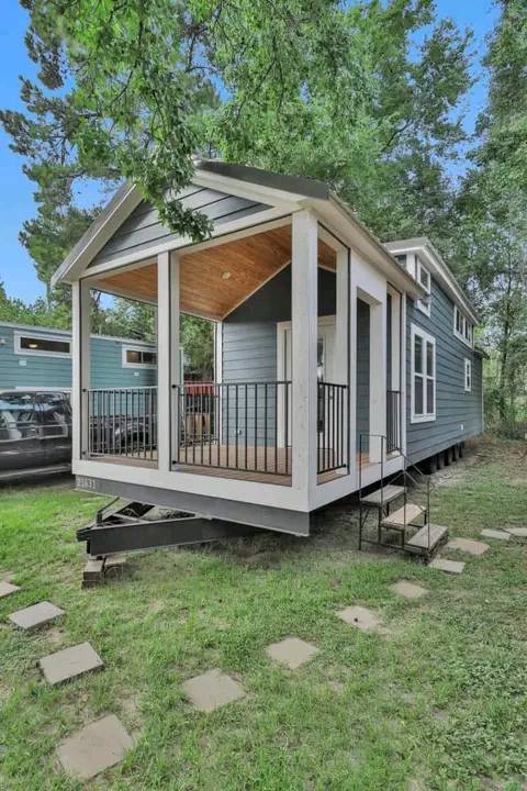 A charming tiny house with blue siding and a white-trimmed porch, set in a lush green area. It features modest steps and a welcoming, cozy atmosphere.