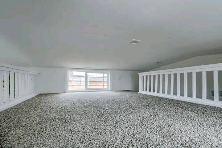 Spacious carpeted loft with low ceiling, white railing, and a large window. The space feels cozy and minimalist, with soft lighting filtering through.