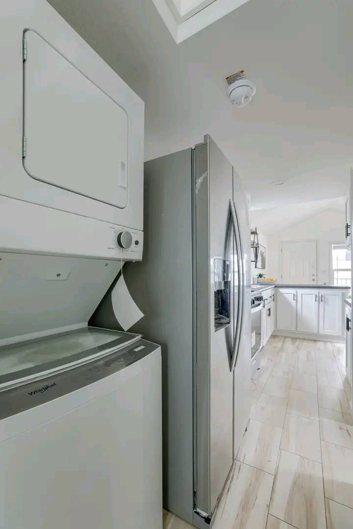 A modern laundry area with a stacked washer and dryer beside a sleek stainless steel refrigerator. The space is bright with white cabinets and light wood flooring, conveying a clean, organized feel.