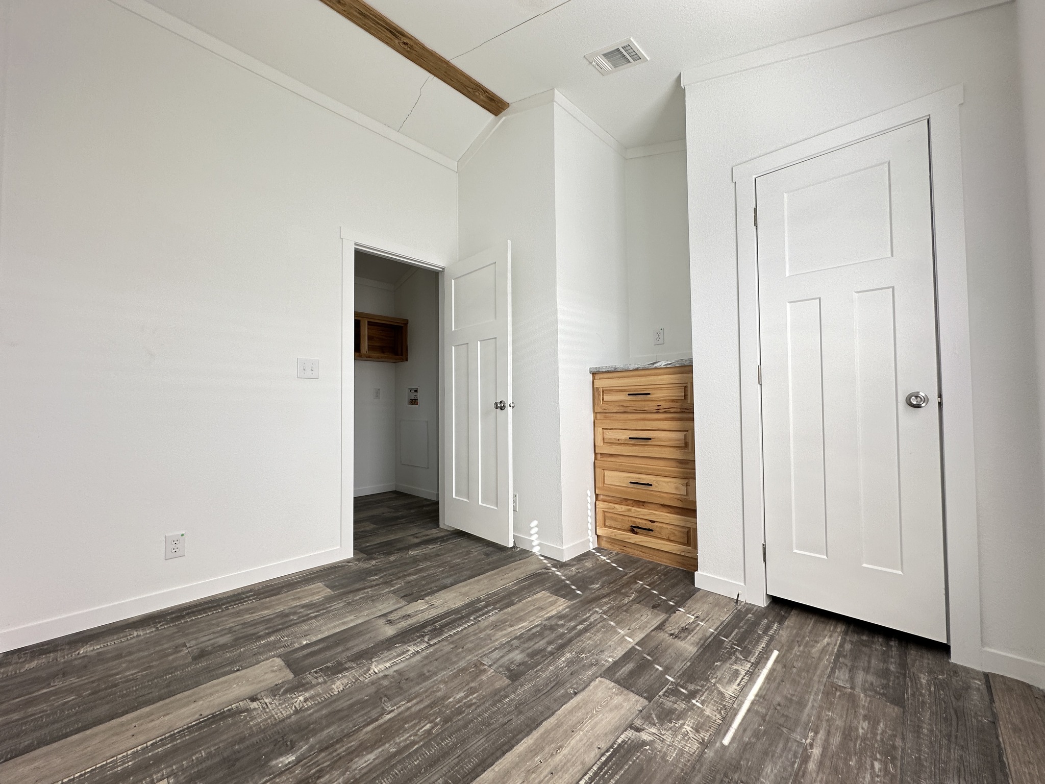 A bright, empty room with white walls, wood-textured flooring, and a ceiling beam. There is a wooden dresser and a door leading to another room.