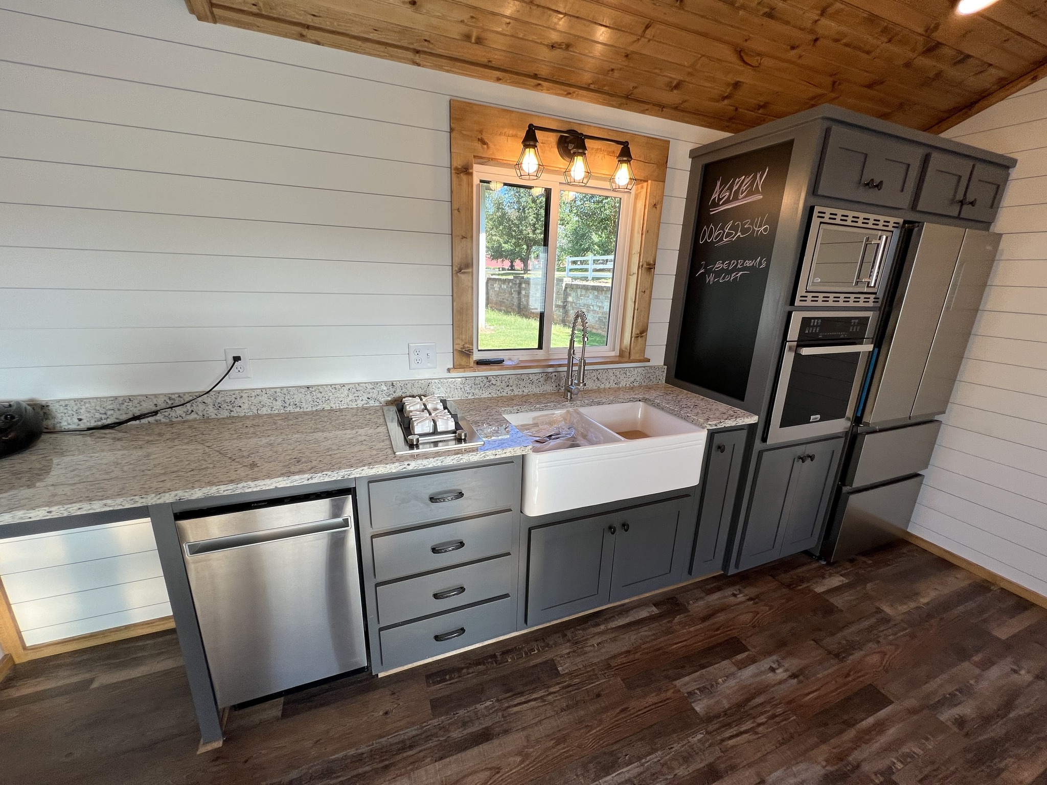 Cozy kitchen with gray cabinets, farmhouse sink, and granite countertops. Window view of outdoors, wooden ceiling, and a fridge with chalkboard notes.
