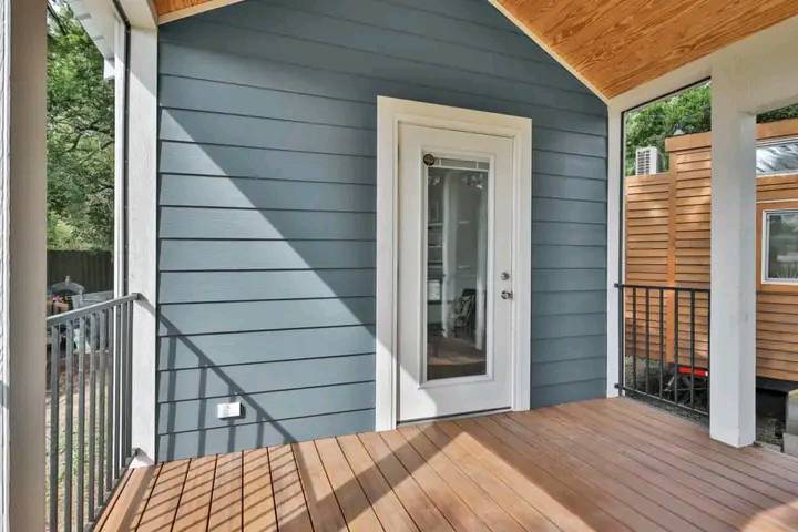 A cozy porch with wooden flooring and blue siding. Sunlight casts shadows, and a glass door leads inside. A calm, inviting outdoor space.