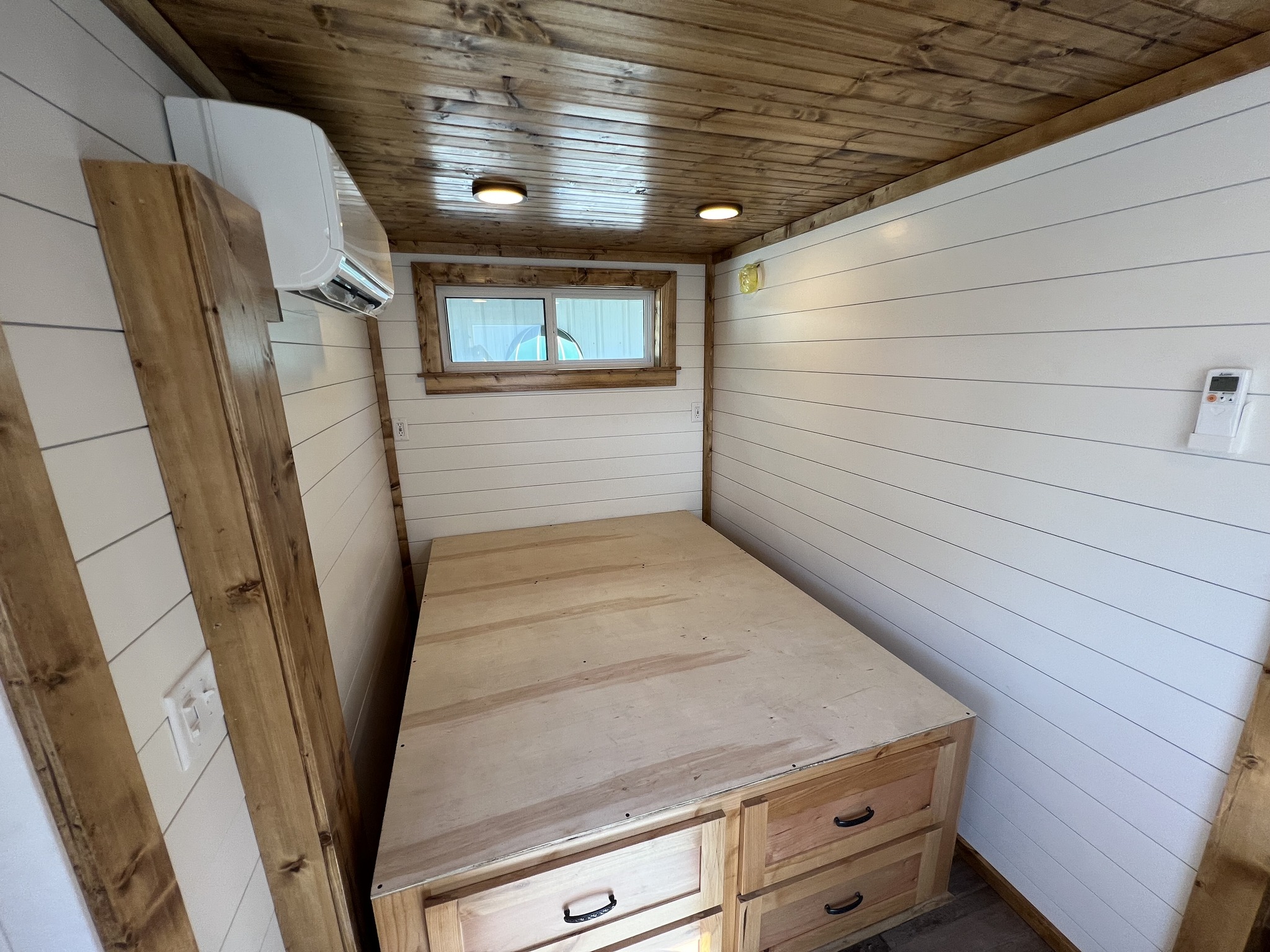 Cozy room with wood paneling, featuring a wooden bed frame and drawers. Soft lighting and a small window create a warm, inviting atmosphere.