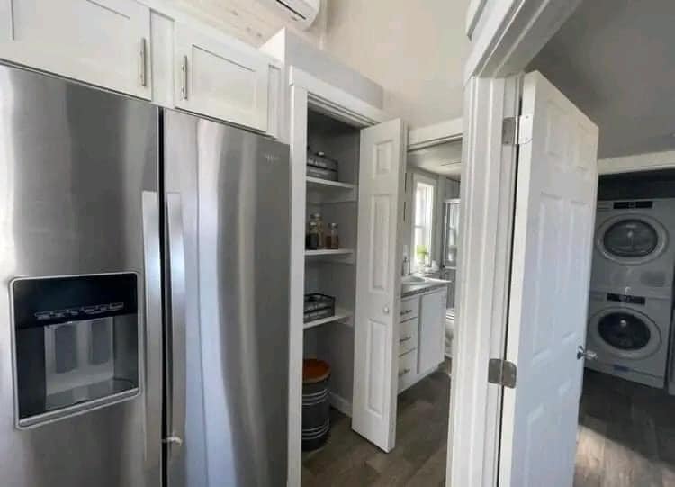 Modern kitchen with stainless steel fridge, open pantry with shelves, and visible laundry area with washer and dryer. The atmosphere is clean and organized.