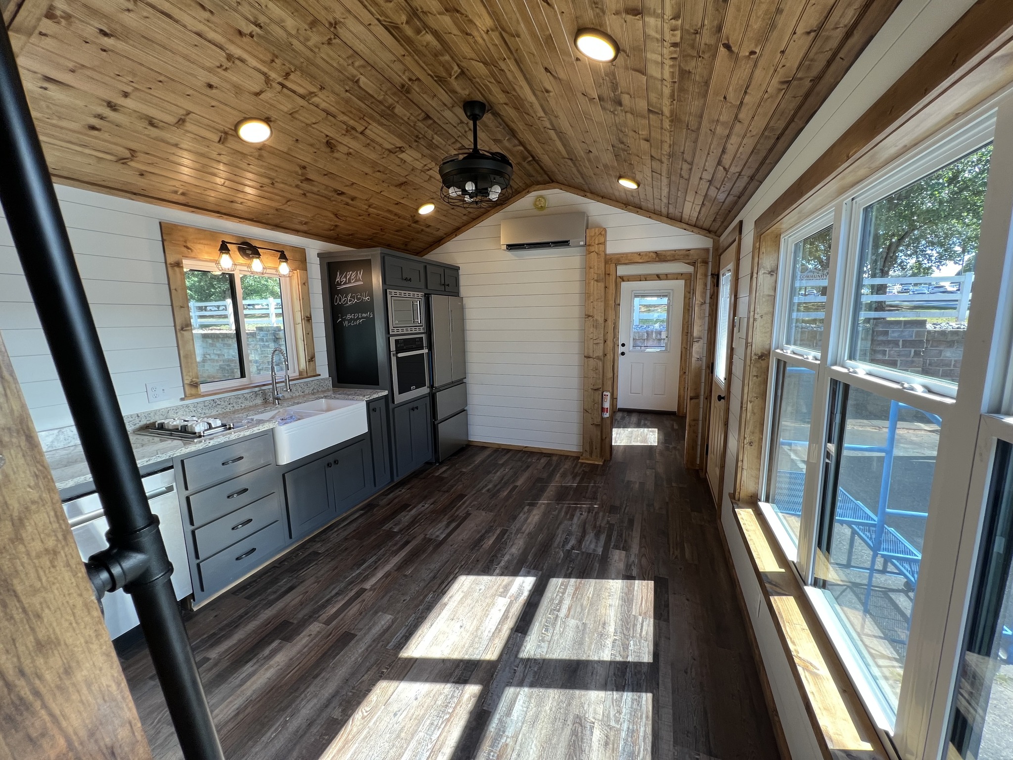 Rustic tiny house kitchen with wood-paneled ceiling and floor, gray cabinets, farmhouse sink, large window, and bright natural light. Cozy and inviting.