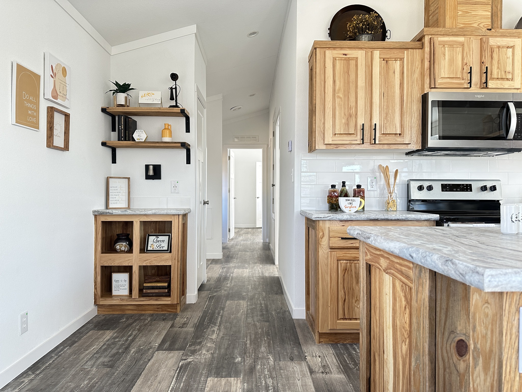 Modern kitchen with natural wood cabinets, rustic shelving, and stainless steel appliances. The hallway leads to a brightly lit room, creating an inviting atmosphere.