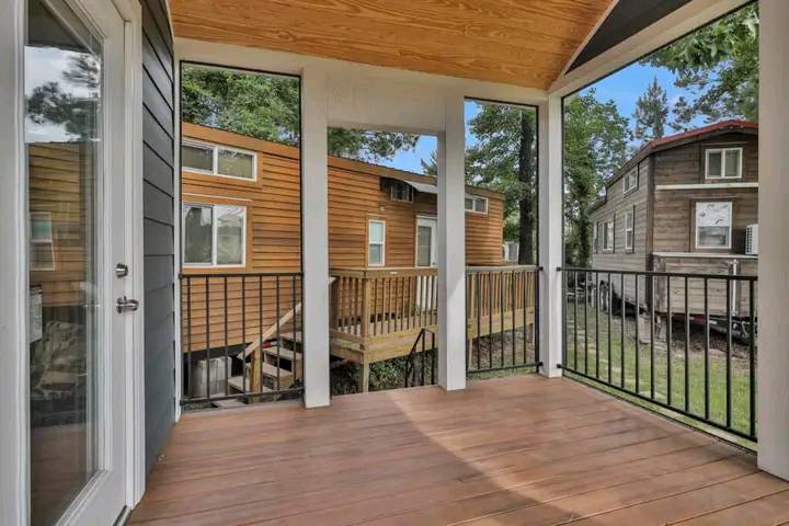 Covered porch with wooden flooring, white columns, and black railing overlooking two brown houses surrounded by trees on a sunny day.