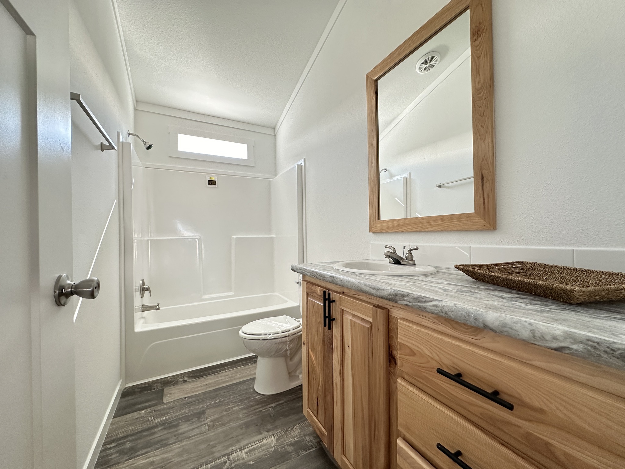 Modern bathroom with a wooden vanity and marble countertop. A large mirror hangs above, and a white bathtub is inset. Natural light from a small window creates a bright, clean atmosphere.