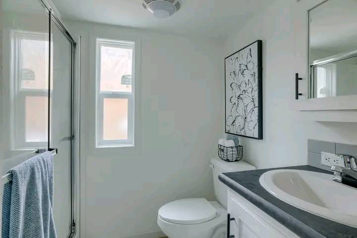 A bright, modern bathroom with white walls and a black countertop. It features a shower, towel, abstract art, and two windows, creating a fresh ambiance.