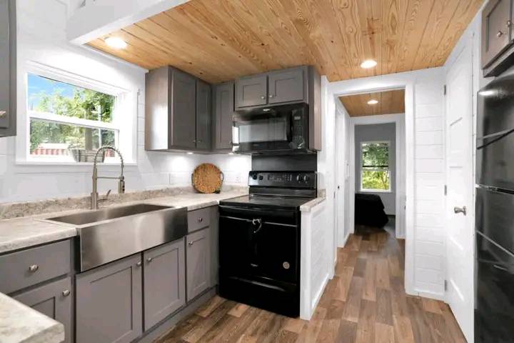 Compact kitchen with gray cabinets and black appliances, featuring a stainless steel farmhouse sink. Wood ceiling and floor add warmth. Natural light from side window.