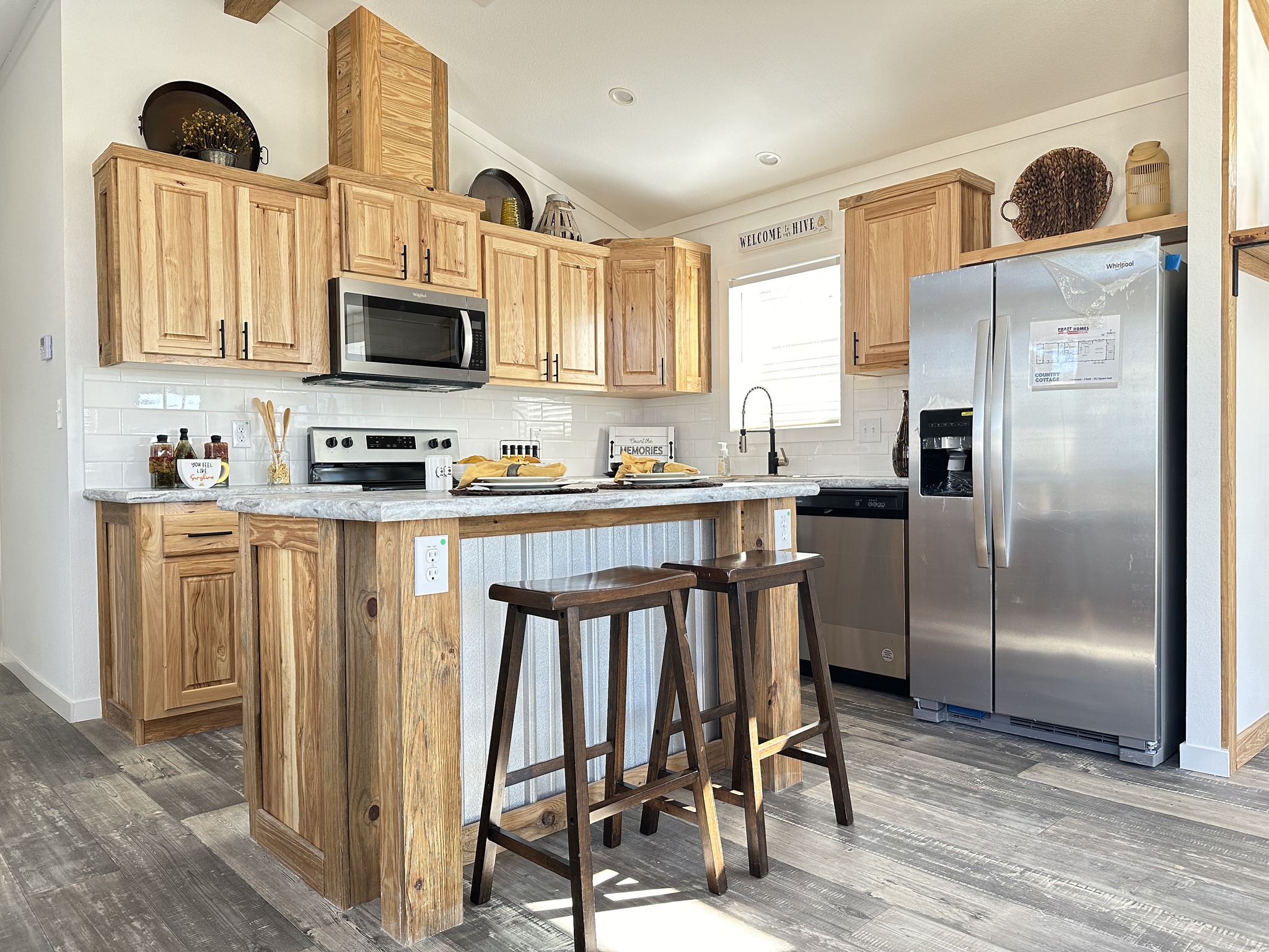 A warm, rustic kitchen features wooden cabinets, a marble island with two stools, stainless steel appliances, and a bright, welcoming ambiance.
