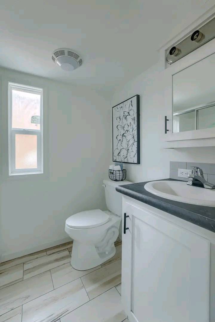 Bright, modern bathroom with white walls and light wood floor. Features a toilet, sink with black countertop, wall art, and a small window for natural light.