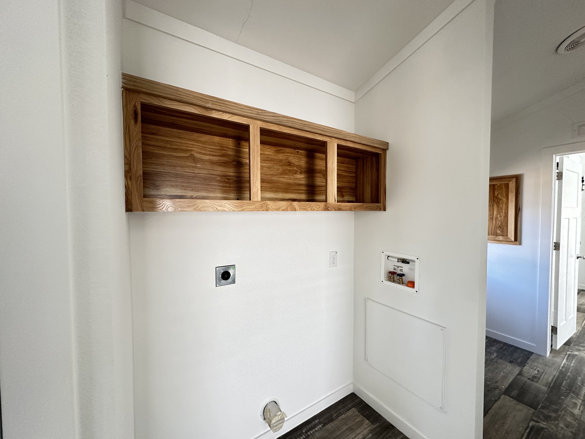 Laundry area with a wooden shelf on a white wall, water hookup and vent openings below. The room has wood flooring and a light, minimalist feel.